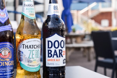 Three bottles of non-alcoholic beverages are arranged on a table in an outdoor setting. The bottles are branded as Erdinger Weissbrau, Thatchers Zero, and Doom Bar Zero. The background features blurred patio furniture and blue umbrellas, suggesting a relaxed and casual environment.