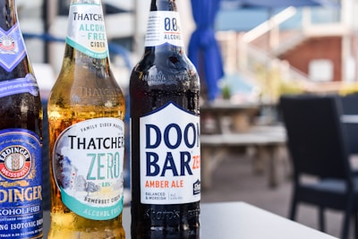 Three bottles of non-alcoholic beverages are arranged on a table in an outdoor setting. The bottles are branded as Erdinger Weissbrau, Thatchers Zero, and Doom Bar Zero. The background features blurred patio furniture and blue umbrellas, suggesting a relaxed and casual environment.
