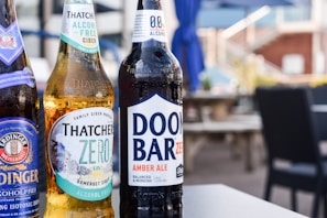 Three bottles of non-alcoholic beverages are arranged on a table in an outdoor setting. The bottles are branded as Erdinger Weissbrau, Thatchers Zero, and Doom Bar Zero. The background features blurred patio furniture and blue umbrellas, suggesting a relaxed and casual environment.