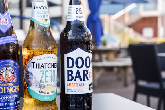 Three bottles of non-alcoholic beverages are arranged on a table in an outdoor setting. The bottles are branded as Erdinger Weissbrau, Thatchers Zero, and Doom Bar Zero. The background features blurred patio furniture and blue umbrellas, suggesting a relaxed and casual environment.