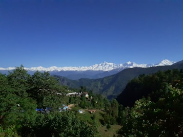 A picturesque landscape featuring a vast expanse of lush green hills in the foreground with a backdrop of towering, snow-capped mountains under a clear, deep blue sky. Small structures are nestled among the greenery, giving a sense of tranquility and seclusion.