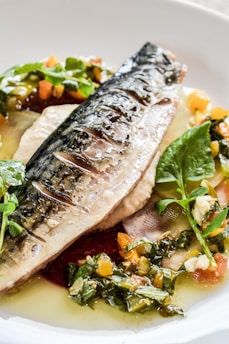 A dish of grilled fish fillet, likely mackerel, served on a creamy base with vibrant greens, diced vegetables, and herbs. The fish has a slightly charred skin showcasing grill marks, and it is garnished with fresh microgreens and chopped vegetables, possibly including bell peppers and leafy greens, all drizzled with a light sauce.