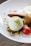 A dessert plate featuring a scoop of pale green ice cream, garnished with finely crushed nuts. A chocolate cookie leans against the ice cream. Fresh strawberries and a creamy panna cotta are also present, with decorative petals scattered around.