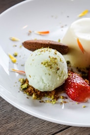A dessert plate featuring a scoop of pale green ice cream, garnished with finely crushed nuts. A chocolate cookie leans against the ice cream. Fresh strawberries and a creamy panna cotta are also present, with decorative petals scattered around.