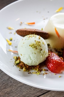 A dessert plate featuring a scoop of pale green ice cream, garnished with finely crushed nuts. A chocolate cookie leans against the ice cream. Fresh strawberries and a creamy panna cotta are also present, with decorative petals scattered around.
