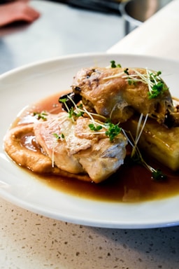 A white plate holds a well-presented dish featuring pieces of cooked chicken garnished with fresh green herbs. The chicken is resting on a bed of sauce, alongside what appears to be a slice of roasted eggplant. The dish is set on a countertop with a subtle terrazzo pattern, suggesting a dining or kitchen environment.
