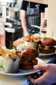 A restaurant setting with a focus on two plates of food featuring burgers and cups of fries. The burgers are served on plates with a garnish of lettuce, and one hand is visible holding a plate. The background shows a blurred figure in a striped apron and kitchen equipment.
