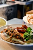 A plate featuring a cooked fish garnished with herbs and a savory sauce, accompanied by sautéed prawns and a side of fresh greens. The meal is presented on a light-colored plate in a softly lit kitchen setting.