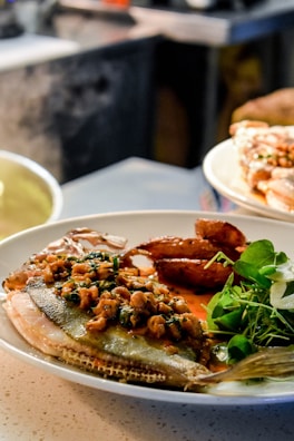 A plate featuring a cooked fish garnished with herbs and a savory sauce, accompanied by sautéed prawns and a side of fresh greens. The meal is presented on a light-colored plate in a softly lit kitchen setting.