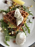 A plate featuring savory waffles topped with a dollop of cream, accompanied by a poached egg and garnished with fresh arugula. Small pieces of corn, beans, and diced vegetables are scattered around, with a lemon wedge on the side.