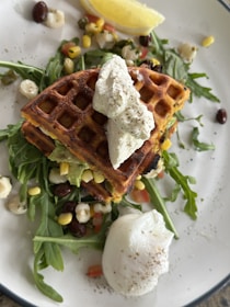 A plate featuring savory waffles topped with a dollop of cream, accompanied by a poached egg and garnished with fresh arugula. Small pieces of corn, beans, and diced vegetables are scattered around, with a lemon wedge on the side.