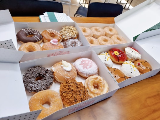 Multiple boxes of assorted donuts are placed on a wooden table. The donuts are of various flavors and toppings, including glazed, chocolate, powdered sugar, cream-filled, and some with colorful decorations. In the background, there are chairs and some notebooks or papers.