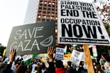 A protest scene with a crowd holding various signs. Prominent signs display messages such as 'Save Gaza' and 'Stand with Palestine! End the Occupation Now!' The atmosphere is filled with activism and solidarity.