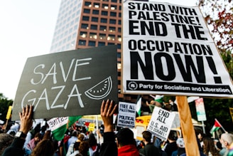 A protest scene with a crowd holding various signs. Prominent signs display messages such as 'Save Gaza' and 'Stand with Palestine! End the Occupation Now!' The atmosphere is filled with activism and solidarity.