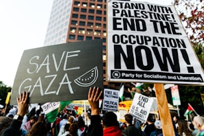 A protest scene with a crowd holding various signs. Prominent signs display messages such as 'Save Gaza' and 'Stand with Palestine! End the Occupation Now!' The atmosphere is filled with activism and solidarity.