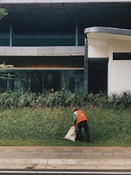 Worker clearing weeds and debris from a garden area near a building.