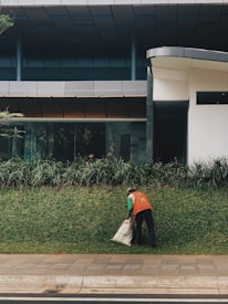 A person wearing an orange vest is cleaning up a grassy area outside a modern building, holding a sack. The area is bordered by a sidewalk, with the building featuring large windows and a sleek design.