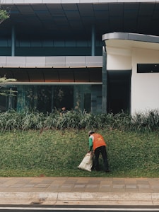 A person wearing an orange vest is cleaning up a grassy area outside a modern building, holding a sack. The area is bordered by a sidewalk, with the building featuring large windows and a sleek design.