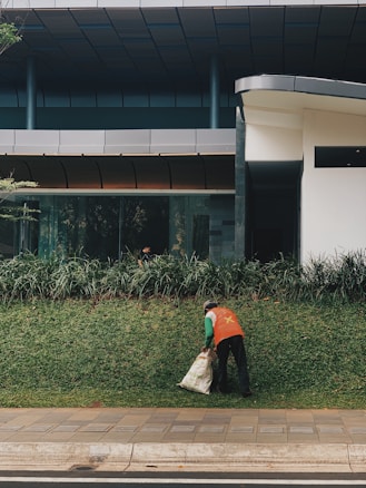 A person wearing an orange vest is cleaning up a grassy area outside a modern building, holding a sack. The area is bordered by a sidewalk, with the building featuring large windows and a sleek design.