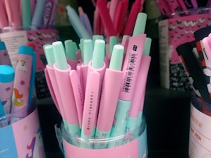 A variety of pastel-colored pens are displayed in a container. The pens are predominantly pink and mint green, with some featuring illustrations of unicorns. The container housing the pens also has pastel designs and unicorn graphics, adding to the whimsical and playful theme.