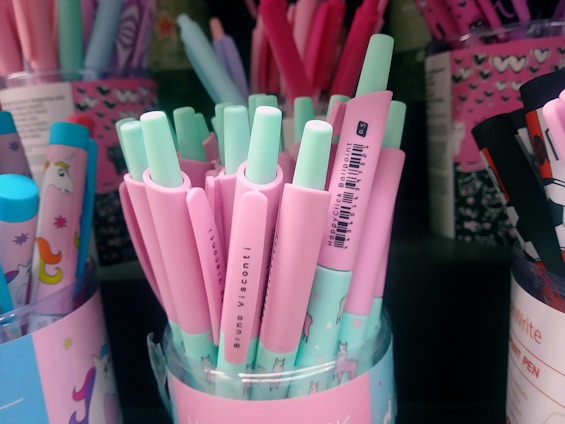 A variety of pastel-colored pens are displayed in a container. The pens are predominantly pink and mint green, with some featuring illustrations of unicorns. The container housing the pens also has pastel designs and unicorn graphics, adding to the whimsical and playful theme.