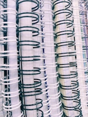 A close-up view of multiple spiral-bound notebooks arranged vertically. The spirals alternate between black and white colors, creating a contrasting pattern. The spines of the notebooks vary in color, including shades of white and light blue.