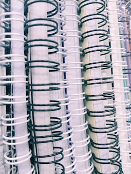 A close-up view of multiple spiral-bound notebooks arranged vertically. The spirals alternate between black and white colors, creating a contrasting pattern. The spines of the notebooks vary in color, including shades of white and light blue.