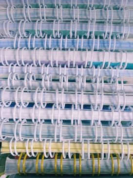 A close-up view of several stacked spiral-bound notebooks, showcasing the binding coils in white and yellow. The pages exhibit a mix of blue, green, and yellow hues.