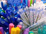 Various writing instruments, including blue and multicolored pens, are clustered together. The pens have clear casings and some feature playful cartoon designs. There are also highlighters in assorted colors such as yellow, pink, and purple.
