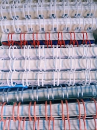 Stacked spiral-bound notebooks with alternating white, red, and blue rings. The notebooks have visible pages with lined paper and a variety of colored covers.