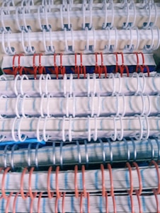 Stacked spiral-bound notebooks with alternating white, red, and blue rings. The notebooks have visible pages with lined paper and a variety of colored covers.
