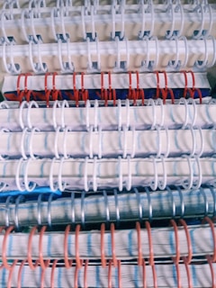 Stacked spiral-bound notebooks with alternating white, red, and blue rings. The notebooks have visible pages with lined paper and a variety of colored covers.