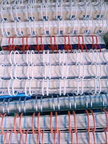 Stacked spiral-bound notebooks with alternating white, red, and blue rings. The notebooks have visible pages with lined paper and a variety of colored covers.