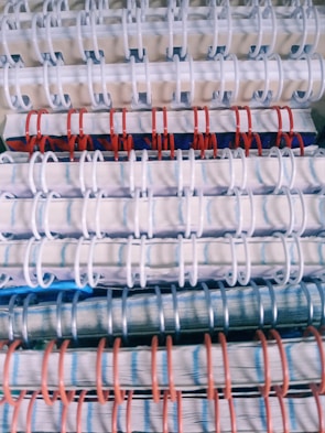 Stacked spiral-bound notebooks with alternating white, red, and blue rings. The notebooks have visible pages with lined paper and a variety of colored covers.