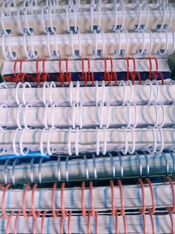 Stacked spiral-bound notebooks with alternating white, red, and blue rings. The notebooks have visible pages with lined paper and a variety of colored covers.