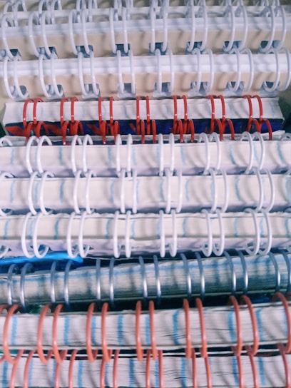 Stacked spiral-bound notebooks with alternating white, red, and blue rings. The notebooks have visible pages with lined paper and a variety of colored covers.