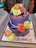 A two-tiered purple cake adorned with colorful icing roses in shades of yellow, orange, and pink. There are multiple candles, some striped, inserted into the cake. The message 'Om Sai Ram' and 'Sai Baba Ji' is written in orange icing on the cake and the platter respectively. The cake is placed in a white box, likely for a celebration or religious occasion.