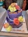 A two-tiered purple cake adorned with colorful icing roses in shades of yellow, orange, and pink. There are multiple candles, some striped, inserted into the cake. The message 'Om Sai Ram' and 'Sai Baba Ji' is written in orange icing on the cake and the platter respectively. The cake is placed in a white box, likely for a celebration or religious occasion.