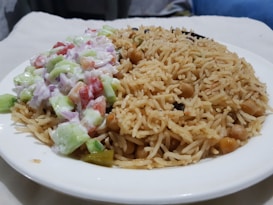 A large white plate contains a serving of rice dish on the right side, featuring long grains and chickpeas, and a mixed vegetable salad on the left with diced cucumbers, tomatoes, and onions in a creamy dressing.