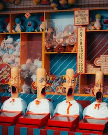 A colorful carnival game booth displays four mannequin heads with exaggerated expressions and open mouths, each wearing a bow tie. Behind them, shelves are filled with various plush toys like blue and red stuffed animals. A sign provides instructions for winning, and there are numbers displayed prominently.