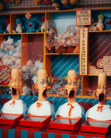 A colorful carnival game booth displays four mannequin heads with exaggerated expressions and open mouths, each wearing a bow tie. Behind them, shelves are filled with various plush toys like blue and red stuffed animals. A sign provides instructions for winning, and there are numbers displayed prominently.