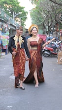 A couple dressed in traditional clothing is walking hand in hand down a street. The man is wearing a black top with golden detailing, a red patterned sarong, and a traditional headdress. The woman is wearing a maroon and gold dress with elaborate patterning, and a headdress adorned with flowers. The street has trees, various shops, and parked motorbikes.