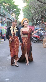 A couple dressed in traditional clothing is walking hand in hand down a street. The man is wearing a black top with golden detailing, a red patterned sarong, and a traditional headdress. The woman is wearing a maroon and gold dress with elaborate patterning, and a headdress adorned with flowers. The street has trees, various shops, and parked motorbikes.