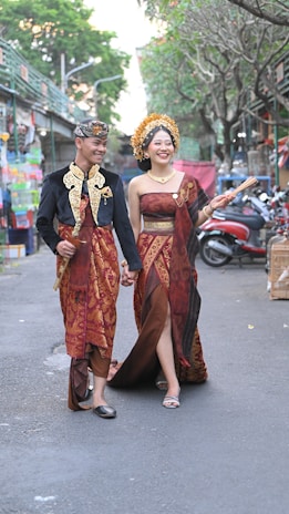 A couple dressed in traditional clothing is walking hand in hand down a street. The man is wearing a black top with golden detailing, a red patterned sarong, and a traditional headdress. The woman is wearing a maroon and gold dress with elaborate patterning, and a headdress adorned with flowers. The street has trees, various shops, and parked motorbikes.