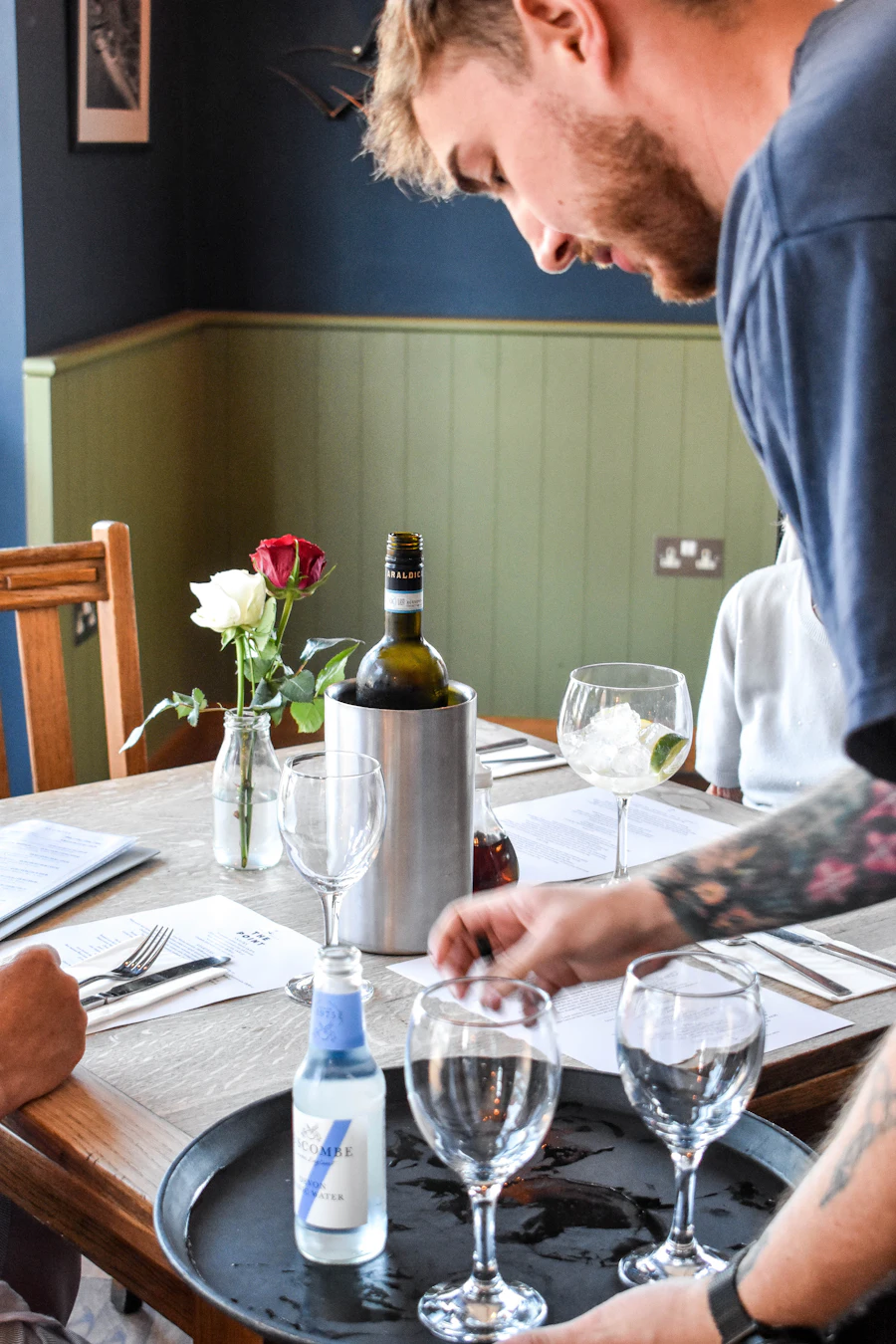 A waiter sets down glasses on a laid table with a wine cooler and a rose.