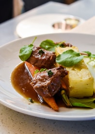 A plate of gourmet food features tender cooked meat garnished with fresh herbs, accompanied by a block of potato gratin and steamed vegetables, all lightly drizzled with a rich brown sauce. The dish is artistically plated and presented on a white dish, sitting on a textured table surface.