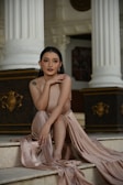 A woman sits gracefully on a set of marble steps, wearing an elegant, flowing, beige gown. Her dark hair is styled neatly, and she appears poised and contemplative. The background features classical architectural elements, including white columns and ornate dark panels with gold accents.