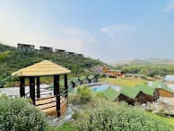 A scenic view of a countryside resort featuring multiple small cabins with green roofs around a central pool. A wooden gazebo with a thatched roof is situated in the forefront, surrounded by lush greenery and flowering plants. The background reveals a hilly landscape with sparse vegetation and several buildings along a ridge.