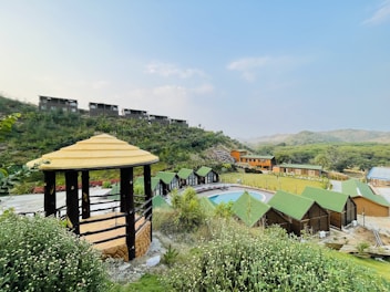 A scenic view of a countryside resort featuring multiple small cabins with green roofs around a central pool. A wooden gazebo with a thatched roof is situated in the forefront, surrounded by lush greenery and flowering plants. The background reveals a hilly landscape with sparse vegetation and several buildings along a ridge.
