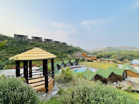 A scenic view of a countryside resort featuring multiple small cabins with green roofs around a central pool. A wooden gazebo with a thatched roof is situated in the forefront, surrounded by lush greenery and flowering plants. The background reveals a hilly landscape with sparse vegetation and several buildings along a ridge.
